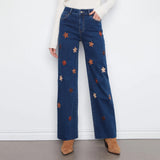 A person wearing dark blue wide-leg jeans with embroidered orange, red, and cream flowers, paired with a white ribbed sweater and tan ankle boots, standing against a white wall.