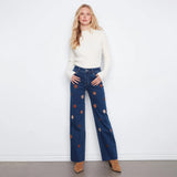 A woman stands against a white wall wearing dark blue wide-leg jeans with embroidered star patterns, a cream ribbed sweater, and tan ankle boots.