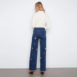 A woman stands with her back to the camera wearing dark blue wide-leg jeans with embroidered star patterns in orange, white, and red, paired with a cream ribbed long-sleeve top.
