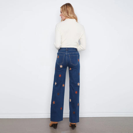 A woman stands with her back to the camera wearing dark blue wide-leg jeans with embroidered star patterns in orange, white, and red, paired with a cream ribbed long-sleeve top.