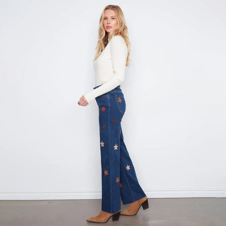 A woman stands in profile wearing blue flare jeans with embroidered star and flower motifs, a white ribbed long-sleeve top, and brown heeled boots against a plain white wall.