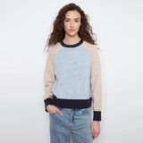 A woman modeling a colorblock raglan sweater with a light blue speckled center, beige sleeves and hem, and dark navy ribbed collar, cuffs, and hem, paired with light-wash blue jeans against a plain white background.