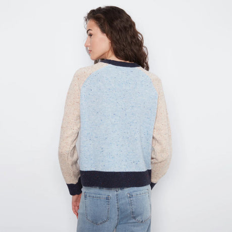 A woman shown from the back wearing a color-block knit sweater with a light blue speckled center panel, beige sleeves, and dark navy ribbed trim, paired with light blue jeans against a plain white background.