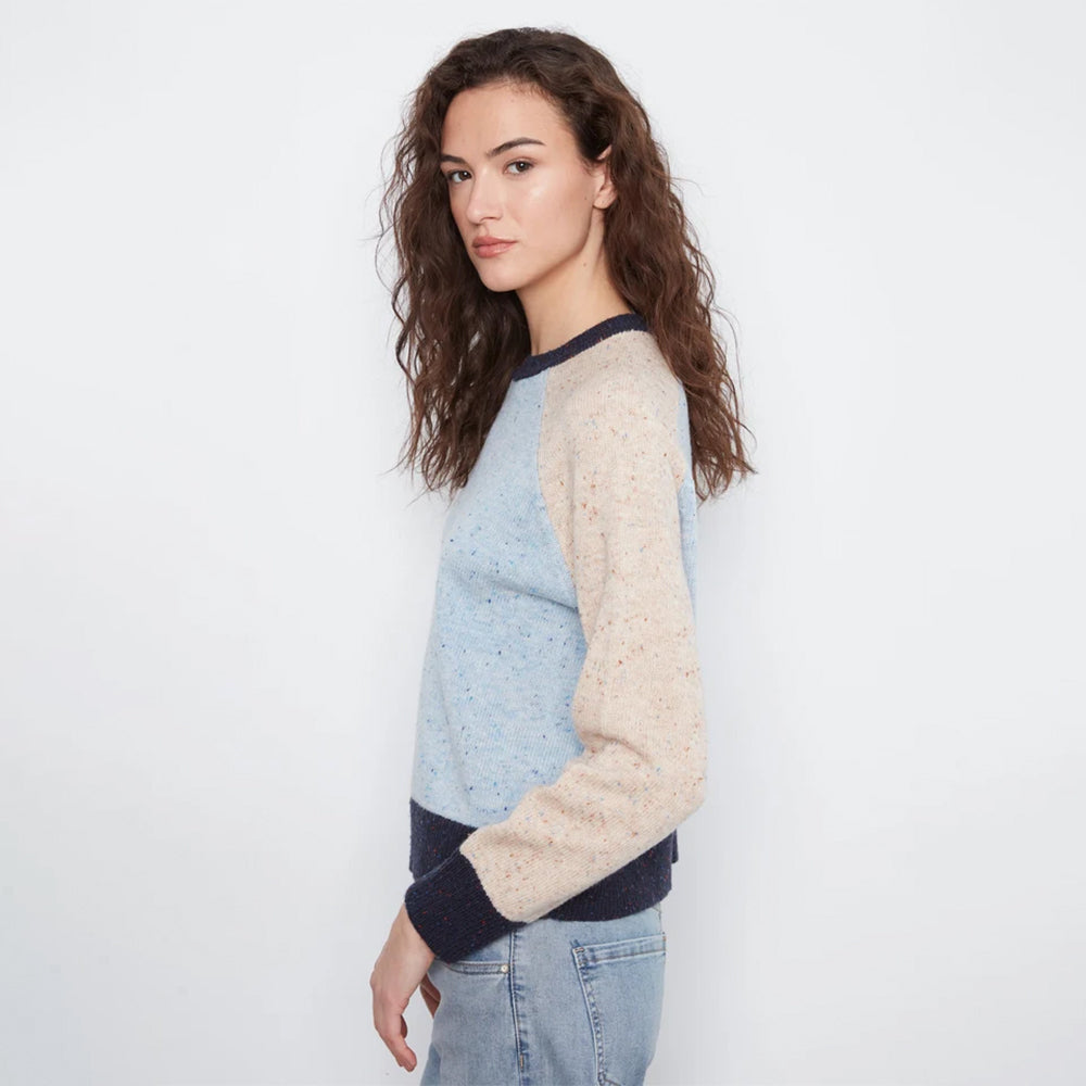 A woman wearing a light blue and beige color-blocked knit sweater with dark navy ribbed trim, paired with light-wash jeans, photographed against a plain white studio background.