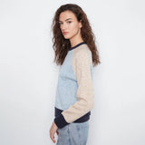 A woman wearing a light blue and beige color-blocked knit sweater with dark navy ribbed trim, paired with light-wash jeans, photographed against a plain white studio background.