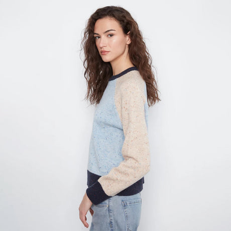 A woman wearing a light blue and beige color-blocked knit sweater with dark navy ribbed trim, paired with light-wash jeans, photographed against a plain white studio background.