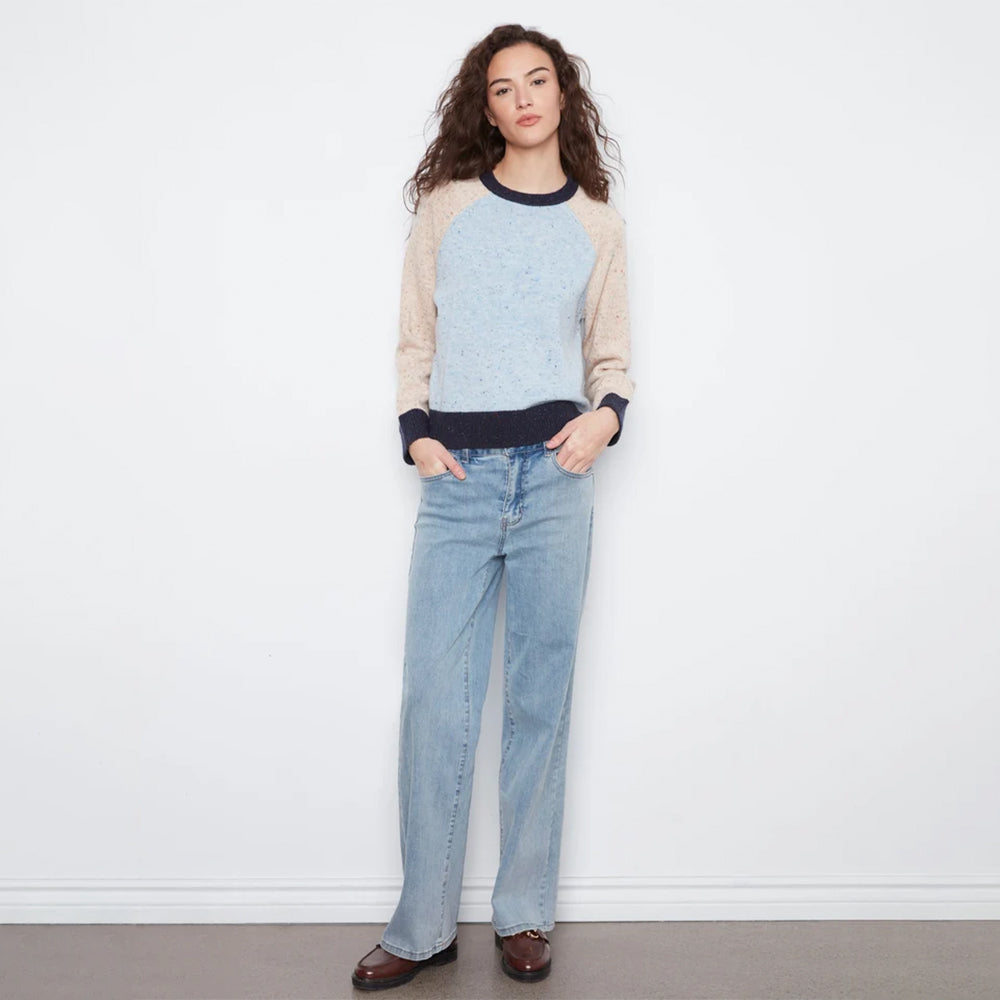 A woman stands against a white wall wearing a light blue and beige raglan sweater with dark navy trim, light-wash blue jeans, and brown leather loafers.
