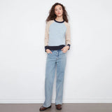 A woman stands against a white wall wearing a light blue and beige raglan sweater with dark navy trim, light-wash blue jeans, and brown leather loafers.