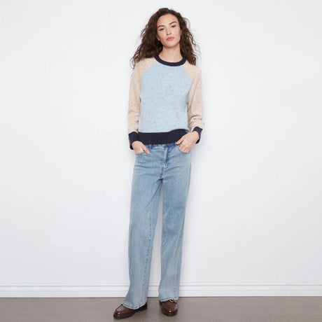 A woman stands against a white wall wearing a light blue and beige raglan sweater with dark navy trim, light-wash blue jeans, and brown leather loafers.