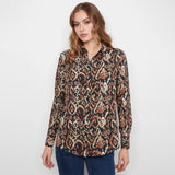 A woman modeling a long-sleeve button-up shirt with an intricate ikat-style pattern in black, beige, and rust orange, paired with blue jeans against a plain white studio background.
