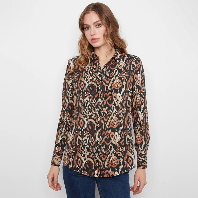 A woman modeling a long-sleeve button-up shirt with an intricate ikat-style pattern in black, beige, and rust orange, paired with blue jeans against a plain white studio background.