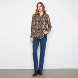 A woman stands against a white wall wearing a long-sleeve button-up shirt with a brown, black, and beige geometric pattern, paired with blue jeans and black ankle boots.
