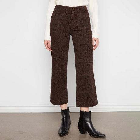 A model wearing dark brown cropped wide-leg trousers with a textured tweed-like pattern, styled with a cream ribbed long-sleeve top and black ankle boots against a white wall and gray floor.