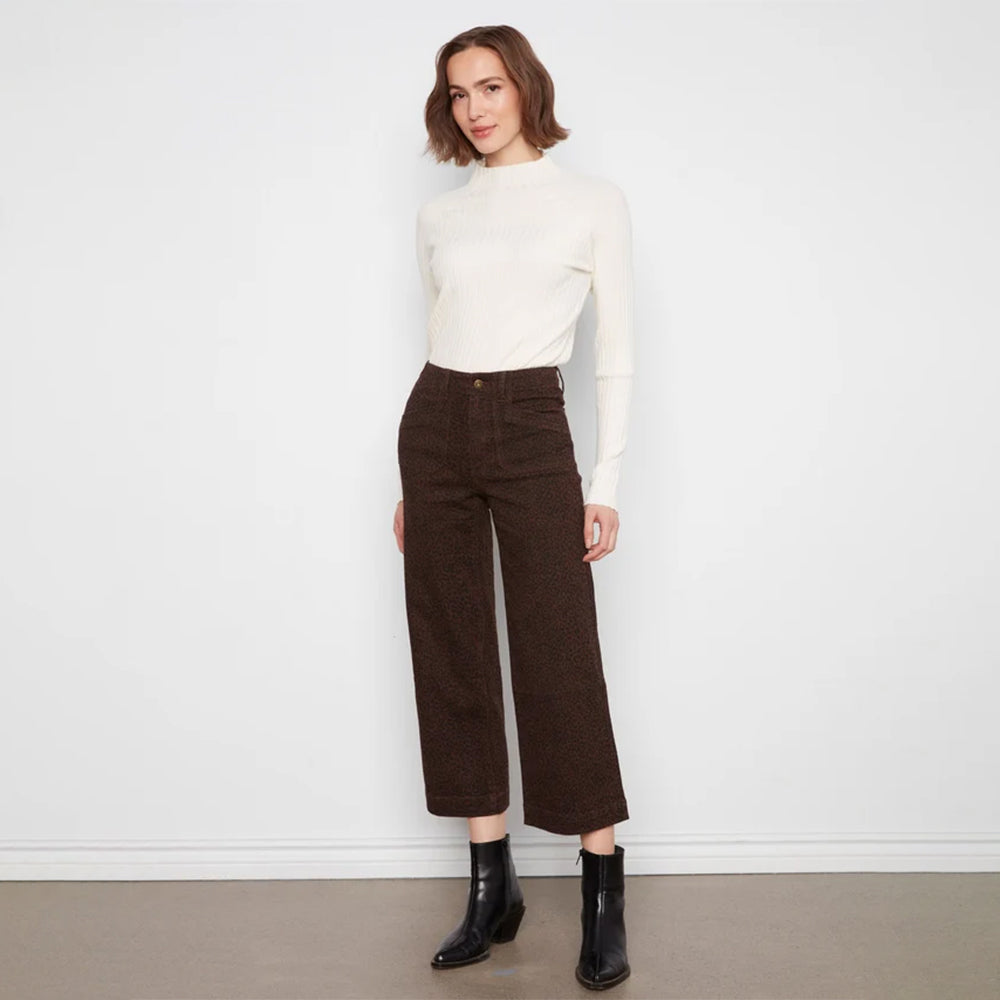 A woman stands against a plain white wall wearing a fitted off-white mock-neck long-sleeve top, dark brown cropped wide-leg trousers, and black ankle boots.