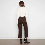 A model viewed from the back wearing dark brown cropped trousers, a cream ribbed turtleneck sweater, and black ankle boots, standing against a plain white wall.