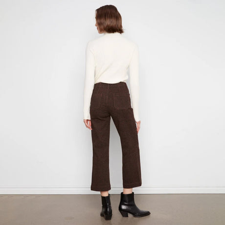 A model viewed from the back wearing dark brown cropped trousers, a cream ribbed turtleneck sweater, and black ankle boots, standing against a plain white wall.