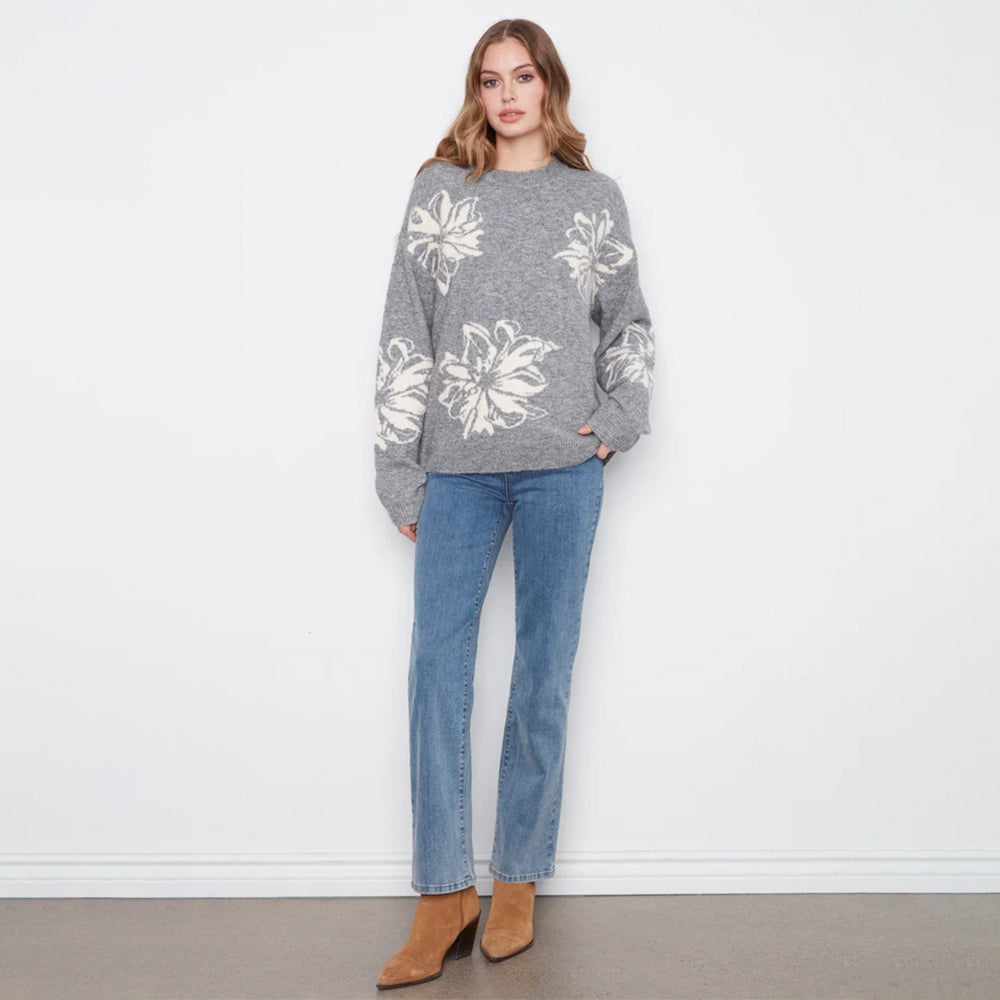 A woman stands against a white wall wearing a gray knit sweater with large white floral motifs, blue jeans, and tan ankle boots.