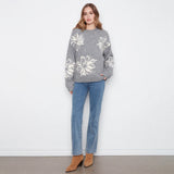 A woman stands against a white wall wearing a gray knit sweater with large white floral motifs, blue jeans, and tan ankle boots.