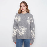 A woman wearing a heather gray knit sweater with large white floral motifs, paired with blue jeans, standing against a plain white studio background.