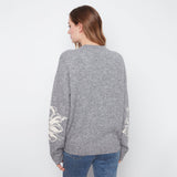 A woman viewed from the back wearing a heather gray knit sweater with cream-colored floral patterns on the sleeves, paired with blue jeans, against a plain white background.