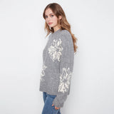 A woman wearing a gray knit sweater with large white floral patterns on the front and sleeves, paired with blue jeans, photographed against a plain white studio background.