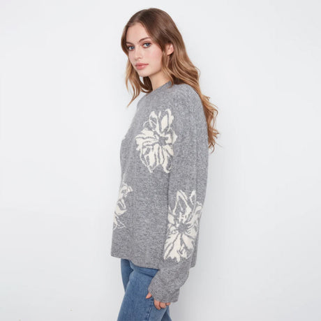 A woman wearing a gray knit sweater with large white floral patterns on the front and sleeves, paired with blue jeans, photographed against a plain white studio background.