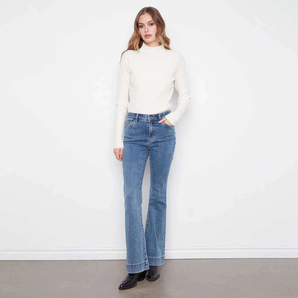 A woman modeling a cream ribbed mock neck sweater and blue high-waisted flare jeans against a white wall and gray floor.
