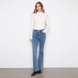 A woman modeling a cream ribbed mock neck sweater and blue high-waisted flare jeans against a white wall and gray floor.