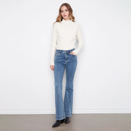 A woman modeling a cream ribbed mock neck sweater and blue high-waisted flare jeans against a white wall and gray floor.