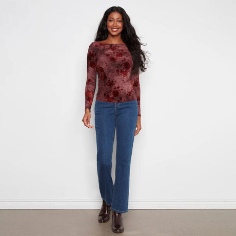 A woman modeling a long-sleeve boat-neck top with a dark red and black floral jacquard pattern, paired with blue jeans and brown ankle boots, standing against a plain white wall.