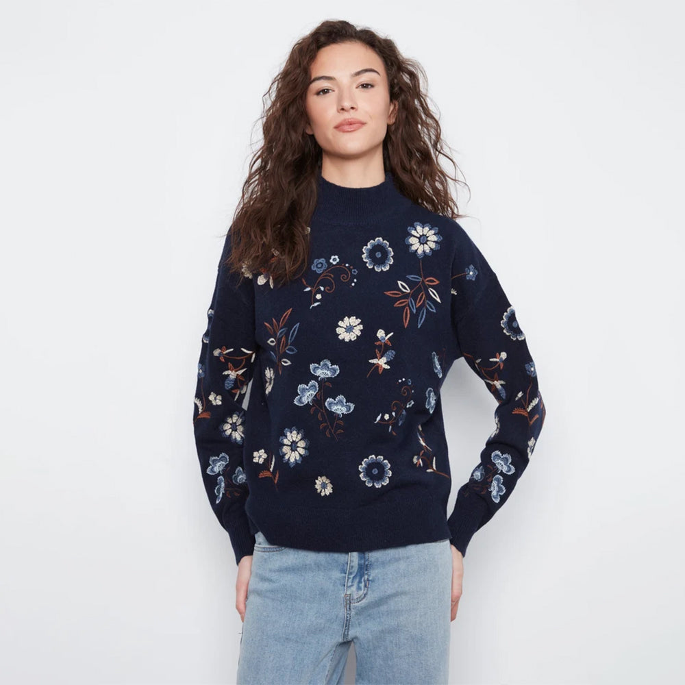 A woman modeling a navy blue knit sweater with a colorful floral pattern, paired with light blue jeans, against a plain white studio background.