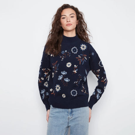 A woman modeling a navy blue knit sweater with a colorful floral pattern, paired with light blue jeans, against a plain white studio background.