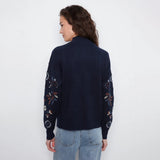 A woman viewed from the back wearing a navy blue knit sweater with floral embroidery on the sleeves, paired with light blue jeans, against a plain white background.