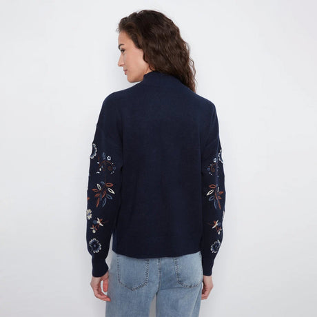 A woman viewed from the back wearing a navy blue knit sweater with floral embroidery on the sleeves, paired with light blue jeans, against a plain white background.