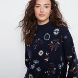 A woman wearing a navy blue mock-neck knit sweater with a delicate embroidered floral pattern in blue, white, and rust orange, set against a plain light gray background.