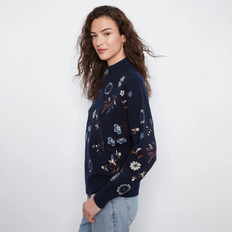 A woman wearing a navy blue knit sweater with a floral pattern, paired with light blue jeans, standing against a plain white studio background.