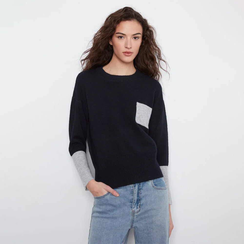 A woman modeling a black knit sweater with light gray contrast panels on the chest and sleeves, paired with light blue jeans, against a plain white studio background.