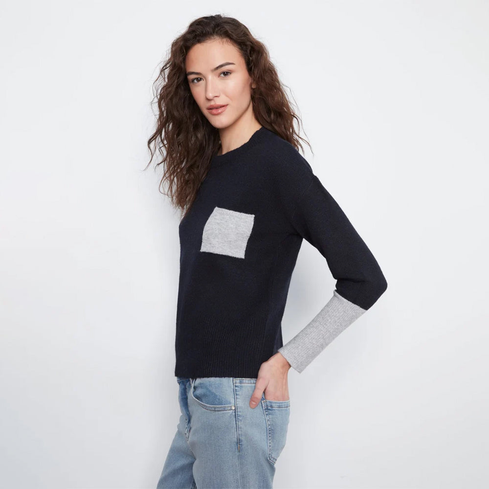 A woman modeling a black long-sleeve knit sweater with a contrasting light gray chest pocket and light gray ribbed cuffs, paired with light blue jeans against a plain white studio background.