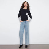 A woman modeling a black long-sleeve knit sweater with a contrasting light gray chest pocket and light gray ribbed cuffs, paired with light blue jeans against a plain white studio background.