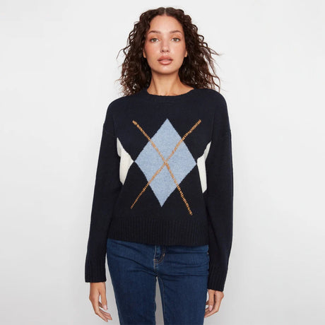 A woman modeling a dark navy blue knit sweater with a light blue, white, and gold argyle pattern on the chest, paired with dark blue jeans against a plain white studio background.