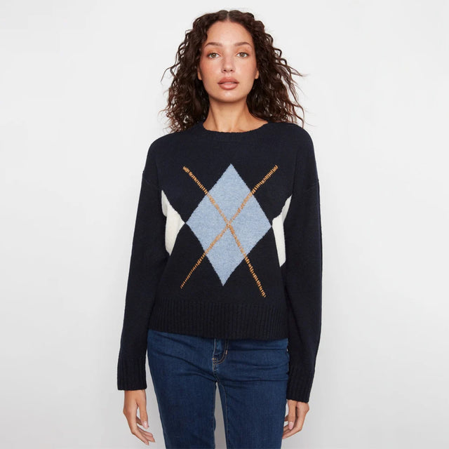 A woman modeling a dark navy blue knit sweater with a light blue, white, and gold argyle pattern on the chest, paired with dark blue jeans against a plain white studio background.