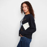 A woman wearing a navy blue knit sweater with a large geometric argyle pattern in white, light blue, and gold, paired with blue jeans, photographed against a plain white studio background.