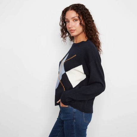 A woman wearing a navy blue knit sweater with a large geometric argyle pattern in white, light blue, and gold, paired with blue jeans, photographed against a plain white studio background.