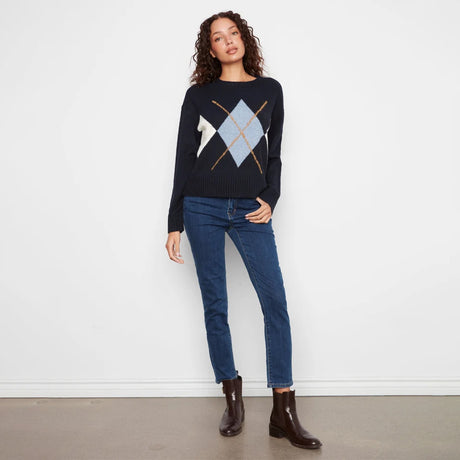 A woman stands against a white wall wearing a dark navy sweater with a light blue and white argyle pattern on the chest, paired with dark blue jeans and dark brown ankle boots.