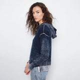 A woman wearing a dark blue and gray acid-wash hooded sweatshirt with white line-art graphics on the shoulders, paired with light-wash blue jeans, photographed against a plain white studio background.