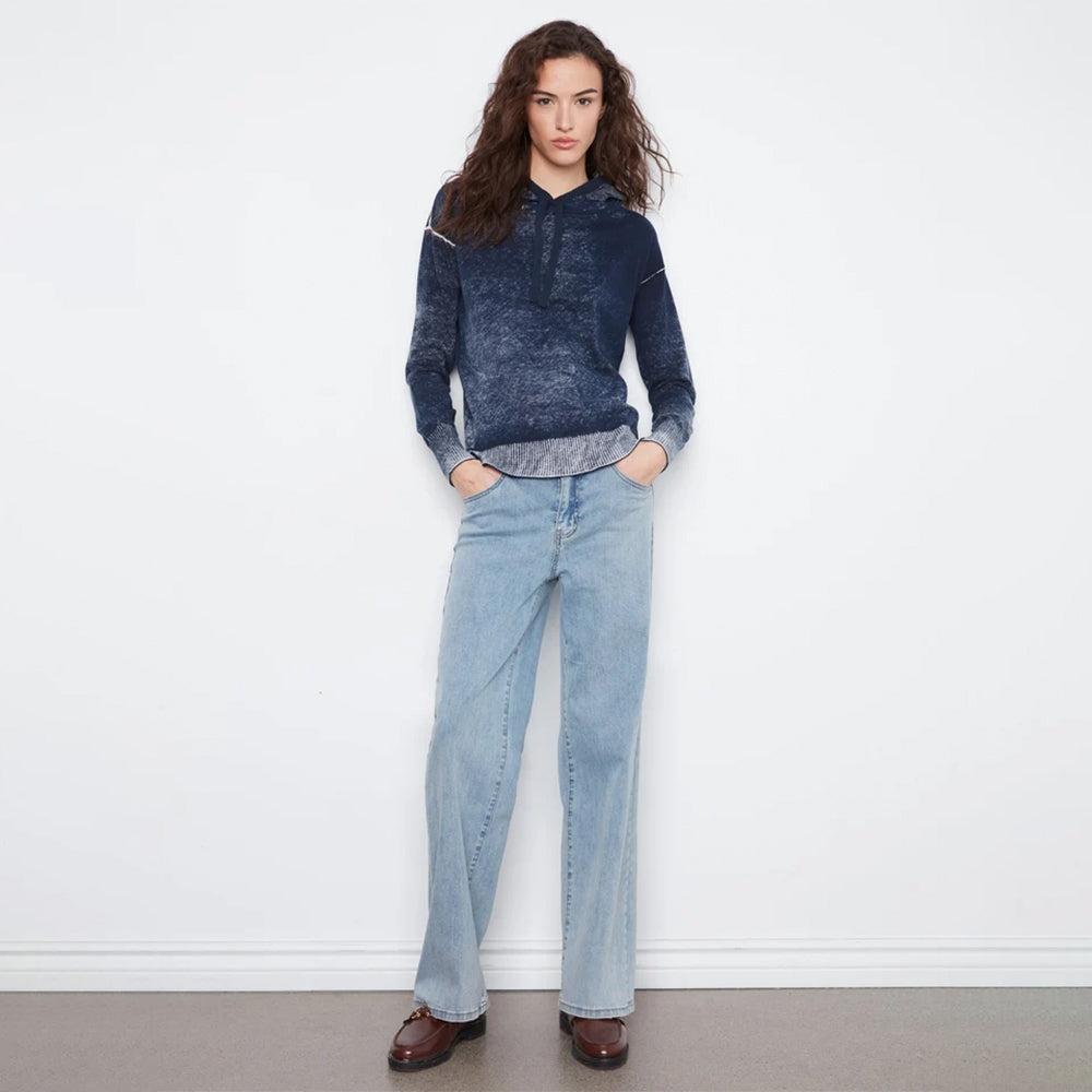 A woman stands against a white wall wearing a dark blue marled knit hoodie with light blue wide-leg jeans and dark brown leather shoes.