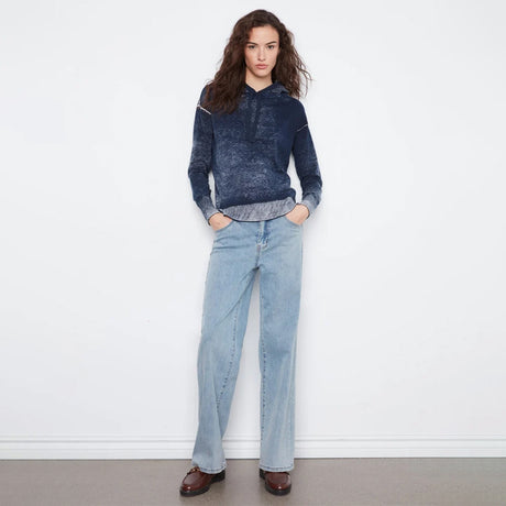 A woman stands against a white wall wearing a dark blue marled knit hoodie with light blue wide-leg jeans and dark brown leather shoes.