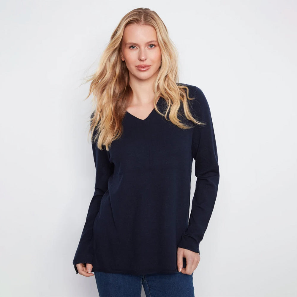 A woman with blonde hair wearing a dark navy blue long-sleeve V-neck top and blue jeans, standing against a plain white studio background.