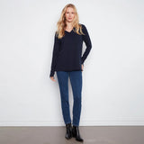 A woman modeling a dark navy blue long-sleeve V-neck knit top, paired with blue jeans and black ankle boots, standing against a plain white wall.