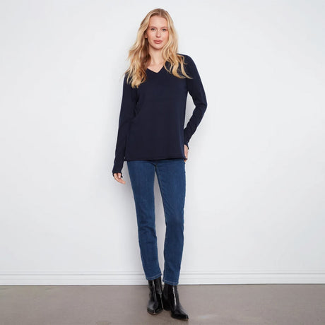 A woman modeling a dark navy blue long-sleeve V-neck knit top, paired with blue jeans and black ankle boots, standing against a plain white wall.